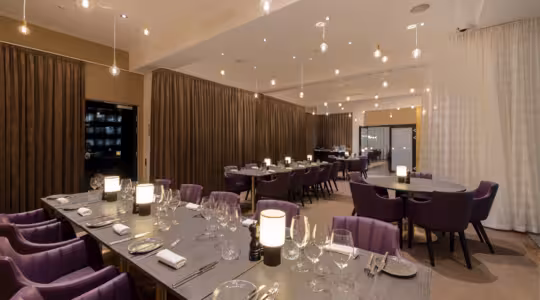 En modern, elegant matsal med lila stolar, träbord med glas, servetter och fat. Mjuk belysning från taklampor skapar en varm atmosfär. Rummet är avdelat av bruna gardiner.