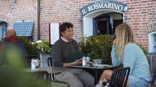 En man och en kvinna sitter vid ett uteserveringsbord, med mannen som håller i en meny. De är engagerade i samtal. En tegelbyggnad med en skylt som säger "Il Rosmarino" finns i bakgrunden. Det finns växter runt sittgruppen.