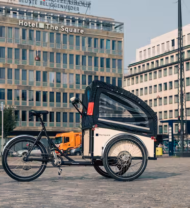 En lastcykel med svart och vit kapell står parkerad på en kullerstensgata i en stad. Bakom det finns moderna byggnader, inklusive ett hotell som heter "The Square", några lastbilar och träd. Himlen är klar.