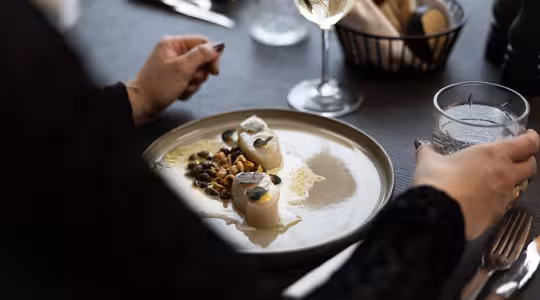 Person som äter vid ett dukat bord med en tallrik pilgrimsmusslor garnerade med örter och nötter. De håller ett glas med is, och det finns ett glas vitt vin och en brödkorg i närheten. Miljön är elegant och svagt upplyst.
