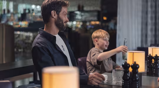En man och en pojke sitter vid ett bord i en svagt upplyst restaurang. Mannen bär en mörk jacka och ler medan han tittar på pojken, som håller i en glasflaska och skrattar. Flera lampor och pepparkvarnar står på bordet.
