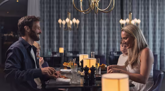 En man och kvinna delar en måltid i en svagt upplyst restaurang med modern inredning och ljuskronor. De sitter vid ett bord med mat och dryck och verkar vara engagerade i konversationer. Två barn syns i bakgrunden.
