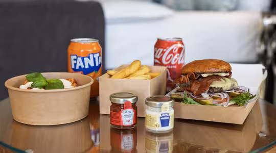 Ett runt glasbord med en cheeseburgare och pommes frites i bruna lådor, en skål pasta med basilika, två små kryddburkar och burkar med Fanta och Coca-Cola i bakgrunden.