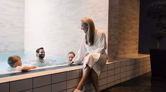 En kvinna i vit morgonrock och tofflor sitter vid kanten av en inomhuspool och ler mot tre personer som är i vattnet. Scenen är varm och avkopplande, med modernt kakel och dämpad belysning som förstärker den mysiga atmosfären.