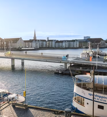 En naturskön utsikt över en hamn i Köpenhamn, med en gångbro med människor som går och cyklar. Båtar lägger till i närheten och historiska byggnader kantar vattnet under en klarblå himmel.