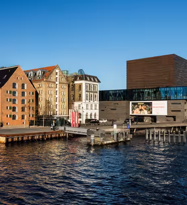 Scen vid vattnet med moderna glasbyggnader och rustika tegelkonstruktioner längs kajen under en klarblå himmel. Dockade båtar syns i förgrunden och reflekterar det lugna vattnet.