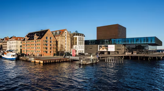 Scen vid vattnet med moderna glasbyggnader och rustika tegelkonstruktioner längs kajen under en klarblå himmel. Dockade båtar syns i förgrunden och reflekterar det lugna vattnet.