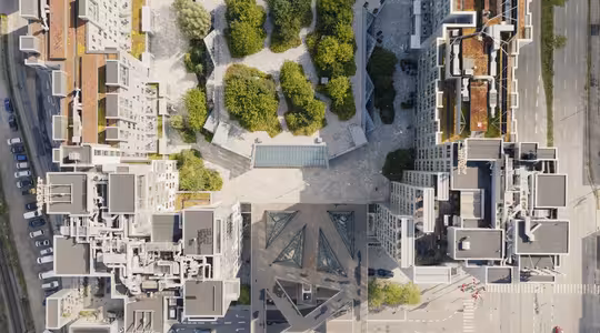Flygfoto över ett stadsområde med en central park med gröna träd och stigar. De omgivande byggnaderna har geometrisk design och modern arkitektur. Gator är synliga i kanterna av bilden.