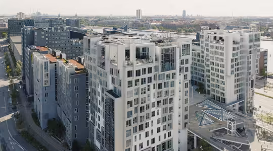 Flygfoto över moderna höghus med stora fönster och balkonger. Skyline bleknar i fjärran. I förgrunden finns en glaspaviljong och omgivande grönska. Himlen är molnig.