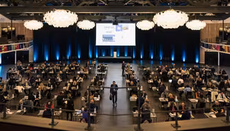 Ett stort konferensrum fyllt med folk vid borden som deltar i ett professionellt evenemang. En skärm visar en timer och presentationsmaterial. Flera ljuskronor hänger i taket och atmosfären är livlig, men ändå organiserad.