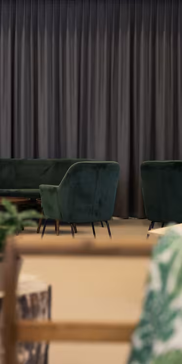 Modern lounge med gröna sammetsfåtöljer, trästolar och små bord. Inredningen inkluderar kuddar med växttema och en vägg med långa, grå gardiner. En varuautomat finns i bakgrunden. Mjuk belysning skapar en mysig atmosfär.
