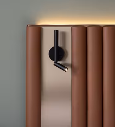 Närbild av en modern, brun stoppad sänggavel mot en ljusgrön vägg. En svart, cylindrisk väggmonterad läslampa är fäst på sänggaveln och kastar ljus uppåt. Sänggaveln har en mjuk, vertikal paneldesign.