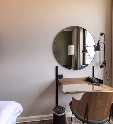 En modern hotellrumshörna har ett träskrivbord med en rund spegel, en svart väggmonterad lampa, en stol, en telefon, en papperskorg och en väggmonterad TV. Beige gardiner täcker delvis ett fönster till höger.