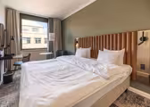Ett modernt hotellrum med en stor säng med vita sängkläder och en brun vadderad sänggavel. Ett skrivbord med en rund spegel är till vänster och ett litet bord med en stol är nära fönstret. Neutrala toner och mjuk belysning skapar en mysig atmosfär.