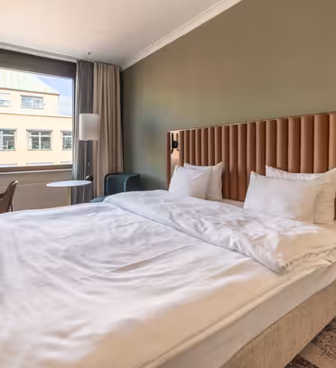 Ett modernt hotellrum med en stor säng med vita sängkläder och en brun vadderad sänggavel. Ett skrivbord med en rund spegel är till vänster och ett litet bord med en stol är nära fönstret. Neutrala toner och mjuk belysning skapar en mysig atmosfär.