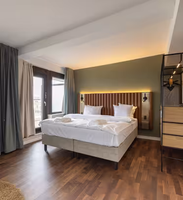 Modernt hotellrum med en snyggt bäddad dubbelsäng, två kuddar och vikta handdukar. Rummet har en grön accentvägg, trägolv, stora fönster med gardiner, en hög lampa, väggmonterad TV och ett litet skåp med hyllor.