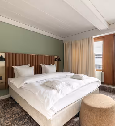 Ett modernt hotellrum med en stor säng med vita sängkläder och sänggavel i trä mot en salviagrön vägg. Det finns en beige ottoman vid fotändan av sängen, och stora fönster med tunga gardiner ger utsikt över en industriell stadsbild.