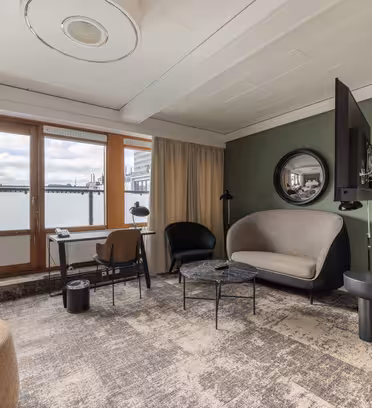 Modernt hotellrum med ett stort fönster och utsikt över balkongen. Har en grå soffa, svart runt bord, snygg stol, golvlampa och väggmonterad TV. Neutraltonade väggar och mattor bidrar till den moderna inredningen.