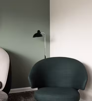 En modern interiör har en beige fåtölj och en mörkgrön stol. En svart, rund spegel hänger på en grön vägg, och bredvid står en svart golvlampa. En vit vägg kontrasterar hörnet av rummet.