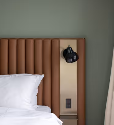 Ett modernt hotellrum med en säng med vita kuddar och lakan. Sänggaveln är brun och vertikalt räfflad. En svart vägglampa är monterad på sänggaveln, bredvid en beige vägg och beige gardiner. Väggen är grönmålad.