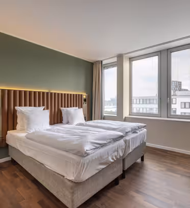 Hotellrum med två snyggt bäddade enkelsängar, en grön accentvägg, trägolv och stora fönster med utsikt över närliggande byggnader. Det finns ett litet runt bord och stoppade stolar vid fönstret. Inredningen är modern och minimalistisk.