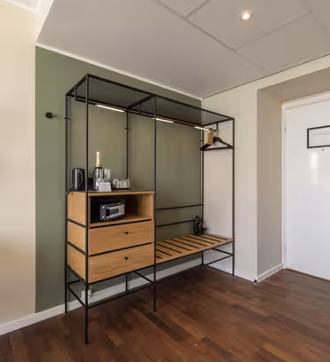 Modern hotellrumsingång med grön accentvägg och öppen garderobsenhet. Skåpet har en champagneflaska, två lådor, vattenkokare och värdeskåp. Väggmonterad lampa och rund spegel ovanför en skrivbordsstol. Vit dörr med elektroniskt lås och trägolv.