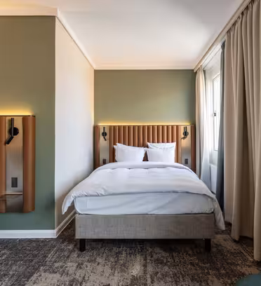 Hotellrum med två enkelsängar, vardera med vita sängkläder och läderklädda sänggavlar. En grön accentvägg är bakom sängarna. Beige gardiner täcker fönstret till höger och en mörkgrön fåtölj syns delvis.