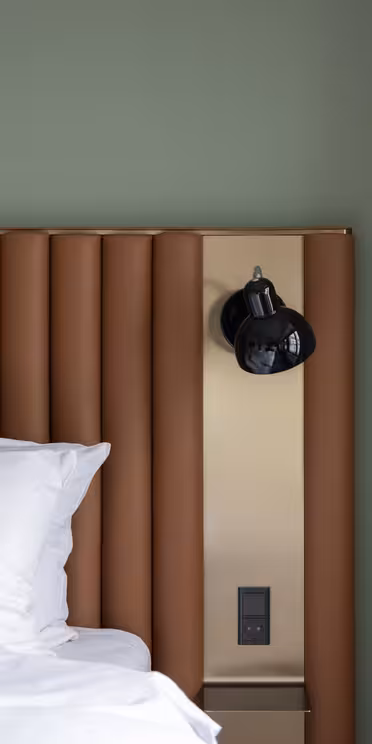 Modernt sovrum med en grön vägg, en brun stoppad sänggavel och en snyggt bäddad säng med vita kuddar och lakan. En svart vägglampa och ett eluttag är monterade på en metallpanel bredvid sängen. Beige gardin till höger.