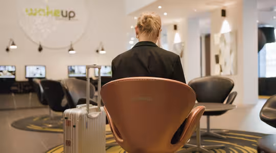 En person med blont hår sitter i en modern lounge, vänd bort från kameran. De har en silver resväska bredvid sig. Utrymmet har en elegant design med stolar, datorer och en skylt som säger wakeup på väggen.