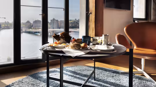 En frukostbricka med bröd, bakverk, ägg, frukt, kaffe och juice står på ett runt bord nära stora fönster med utsikt över stadsbilden vid vattnet. Solljus lyser upp det mysiga rummet med en matta och en orange fåtölj.
