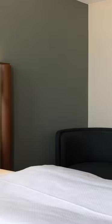 Ett modernt hotellrum med en säng med vita sängkläder och kuddar, en vadderad brun sänggavel, ett litet runt sidobord, en svart fåtölj och en vägghängd lampa med vit skärm.