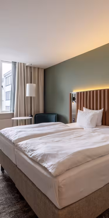 Ett modernt hotellrum med en stor säng, två vita kuddar, en grön accentvägg, väggmonterade lampor, ett skrivbord med en stol, en rund spegel, golvlampa, fåtölj och ett stort fönster med beige gardiner.