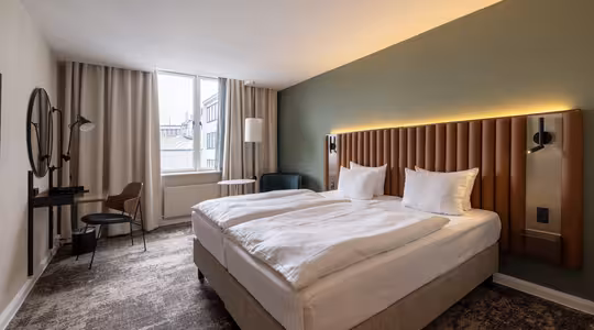 Ett modernt hotellrum med en stor säng, två vita kuddar, en grön accentvägg, väggmonterade lampor, ett skrivbord med en stol, en rund spegel, golvlampa, fåtölj och ett stort fönster med beige gardiner.