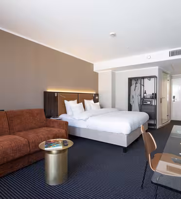 Modernt hotellrum med dubbelsäng, brun soffa, litet runt bord, platt-TV, skrivbord med stol, lampa och öppen garderob. Rummet har neutral inredning och gott om naturligt ljus.