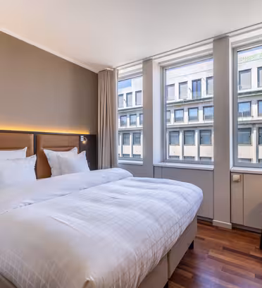 Ett modernt hotellrum med en stor dubbelsäng, vita sängkläder, två fönster med beige gardiner, en brun accentvägg, en brun stoppad fåtölj och en golvlampa bredvid sängen.