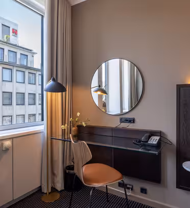 Ett modernt hotellrumshörn med ett glasskrivbord, en brun stol, en rund spegel, en skrivbordslampa, en telefon och en liten vas. Stora fönster släpper in naturligt ljus, och en säng med ett vägghängt nattduksbord är delvis synlig.