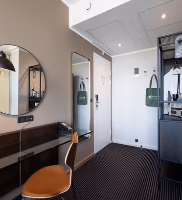 Ett modernt hotellrum med skrivbord, brun stol, rund spegel, svart lampa, hög spegel, klädhängare och en svart hylla med minibar och kaffebryggare mot neutrala väggar och mörk heltäckningsmatta.