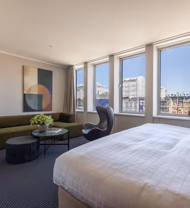 Modernt hotellrum med stora fönster, en king size-säng, en grön soffa, en svart fåtölj, ett litet runt bord med en krukväxt, abstrakt väggkonst och en platt-TV, med utsikt över stadens byggnader utanför.