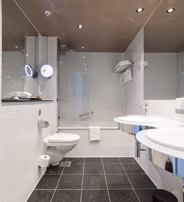 Modernt hotellbadrum med vitkaklade väggar, svart kaklat golv, två runda glashandfat med speglar, en vägghängd toalett, handdukshängare och ett badkar med glasvägg och monterat duschmunstycke.