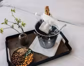 På en bricka på en säng står en vas med gröna blommor, en skål med olika nötter och en ishink av metall med en champagneflaska, två glas och en vikt vit servett.