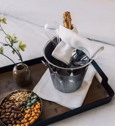 På en bricka på en säng står en vas med gröna blommor, en skål med olika nötter och en ishink av metall med en champagneflaska, två glas och en vikt vit servett.