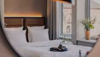 Ett mysigt hotellrum med prydligt bäddade enkelsängar, vita sängkläder, en bricka med frukostartiklar, stora fönster med gardiner och en vas med gröna växter i fönsterbrädan.