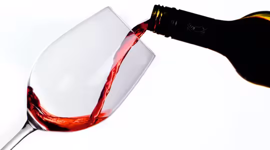 En flaska häller upp rött vin i ett lutande, klart vinglas mot en vit bakgrund. Vinet bildar en jämn, bågande ström när det rinner in i glaset.