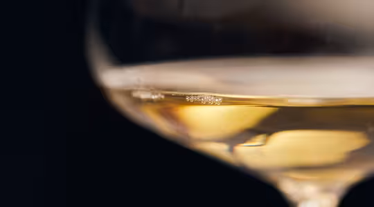 En närbild av ett glas vitt vin, delvis fyllt, med mjukt fokus på den gyllene vätskan inuti. Bakgrunden är mörk och framhäver vinets känsliga färg och de subtila reflektionerna på glasytan.