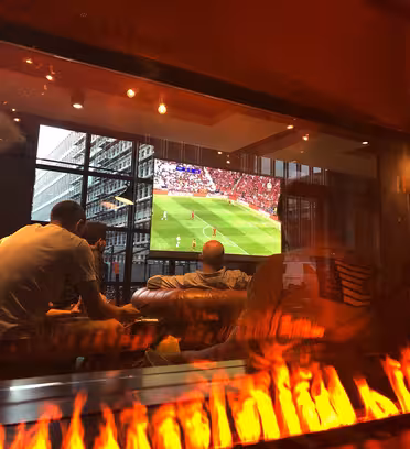 Människor tittar på en fotbollsmatch på en stor skärm inomhus, sedd genom ett glas med reflektioner. En synlig öppen spis förhöjer den mysiga atmosfären. Utanför syns kontor genom fönstren.