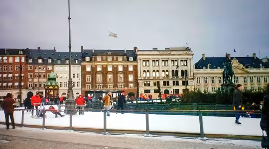 Människor åker skridskor på en rink på ett stadstorg med historiska byggnader i bakgrunden. Scenen innehåller en staty och myllrar av skridskoåkare och åskådare omgiven av en vintrig atmosfär.