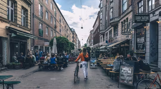 En person som går med en cykel på en livlig gata kantad av tegelbyggnader och uteserveringar. Folk sitter och umgås vid borden. Himlen är delvis molnig och skyltar och grönska pryder området.