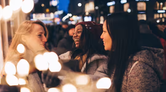 Tre kvinnor ler och pratar i en festlig, upplyst gatumiljö. Scenen är livlig, med suddiga ljus som skapar en varm glöd. Stämningen är glad och livlig med kvällsaktivitet.