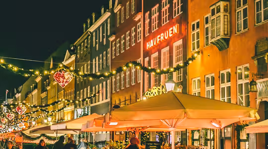 En festlig gatubild på natten, med människor som går förbi restauranger prydda med ljusslingor och upplysta hjärtformade dekorationer. Den varma glöden reflekteras från kullerstensbeläggningen och skapar en mysig atmosfär.