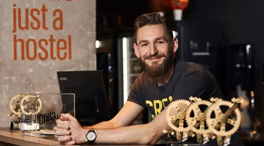 En leende man med skägg lutar sig mot en reception på ett vandrarhem. Han bär en mörk t-shirt och en klocka. Bredvid honom finns flera kuggformade troféer. På väggen bakom honom står det "vi är bara ett vandrarhem" med orange bokstäver.